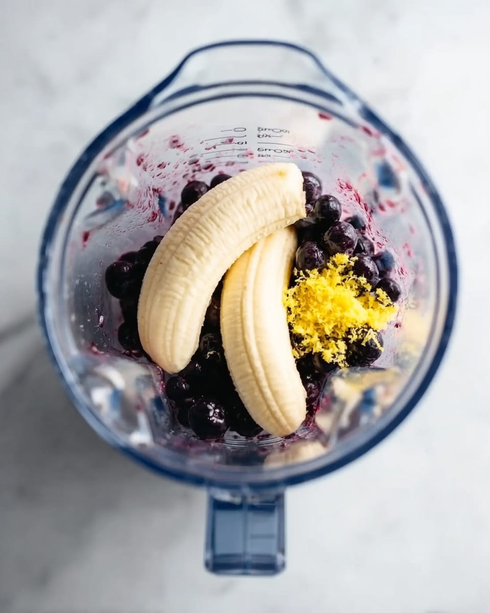 A clear blender jar is shown from the top, resting on a white marbled surface. Inside the blender, there are two peeled bananas placed on top of a layer of dark purple blueberries. There is a small amount of bright yellow grated ingredient, likely lemon zest, scattered to the right side of the bananas and blueberries. The blender jar has clear measurement markings visible on the side. Photo taken with an iphone --ar 4:5 --v 7