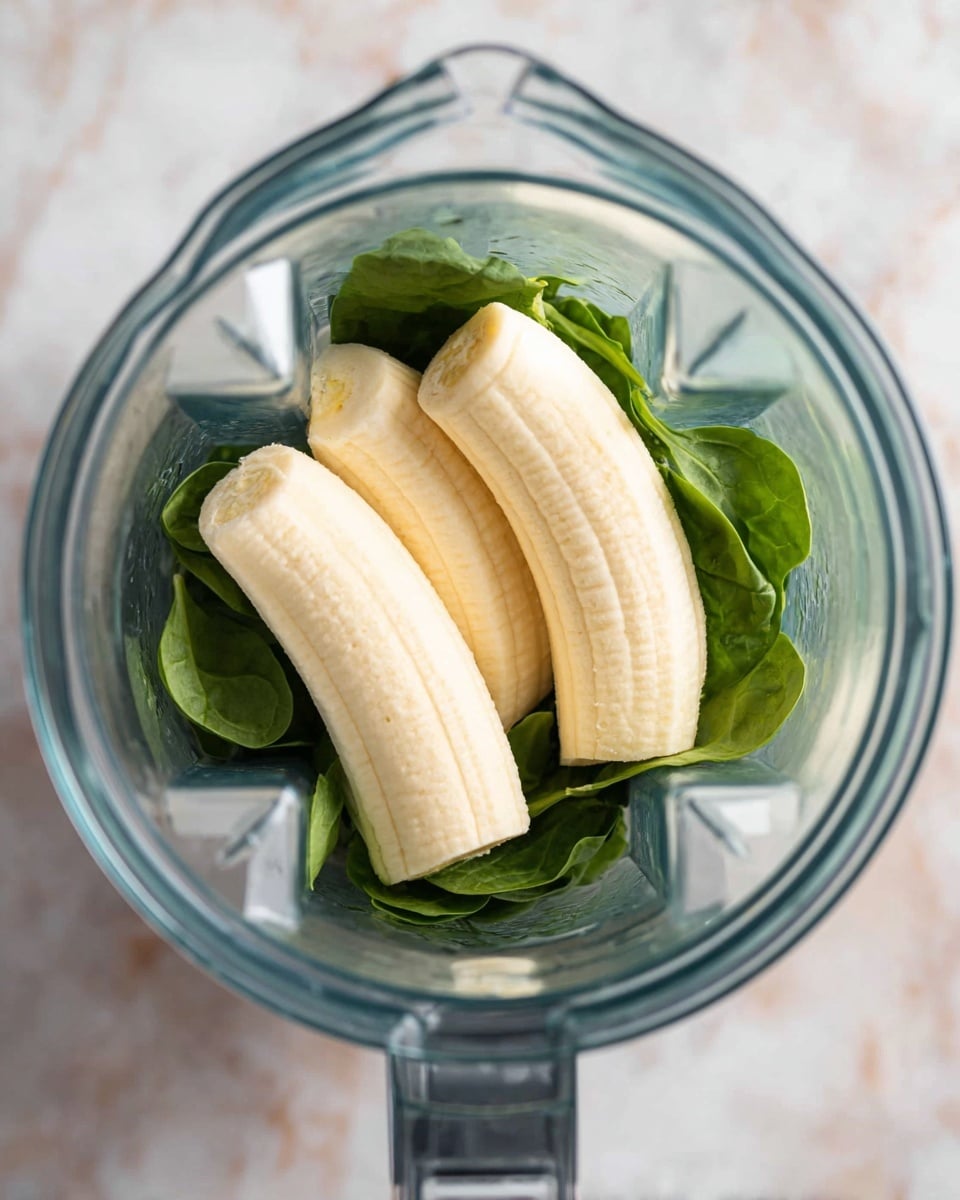 A clear blender jar with three peeled bananas placed on top of a small bunch of fresh spinach leaves. The bananas are light yellow and smooth, lying horizontally in the center. The spinach leaves underneath are dark green with a fresh, slightly wrinkled texture, mostly covering the bottom of the blender. The blender sits on a white marbled surface, shown from above. Photo taken with an iphone --ar 4:5 --v 7