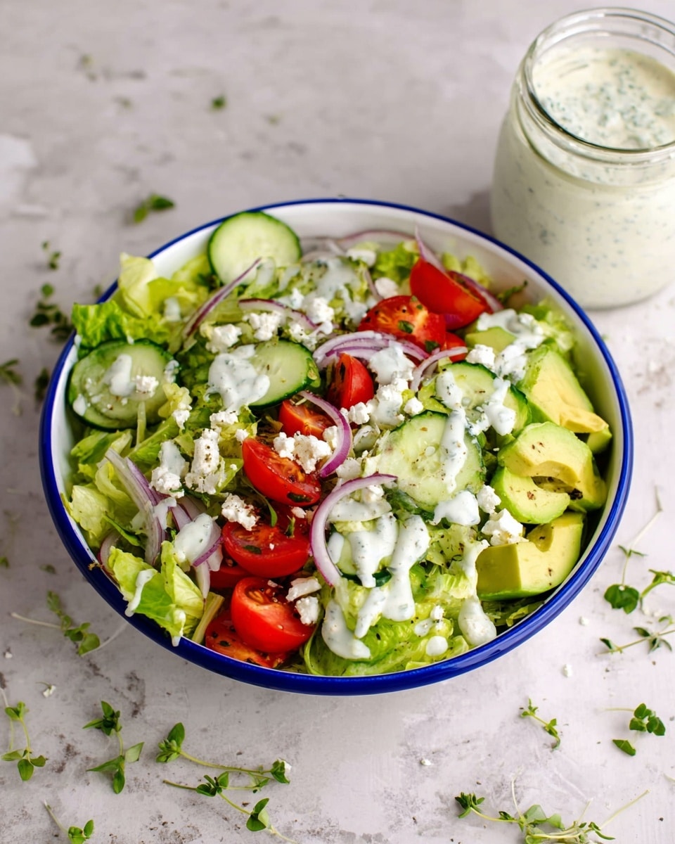 A white bowl with a blue rim is filled with fresh salad layers starting with a base of leafy green lettuce. On top, there are slices of cucumber with light green skin, quartered red cherry tomatoes, and thin rings of red onion. Small chunks of light yellow avocado are scattered throughout. White crumbled cheese pieces and a creamy white dressing drizzled over the salad add texture and contrast. Bits of fresh green herbs are sprinkled around on the salad and outside the bowl on a white marbled surface. In the background, there is a clear glass jar with a white creamy dressing and small green herb pieces on top. photo taken with an iphone --ar 4:5 --v 7
