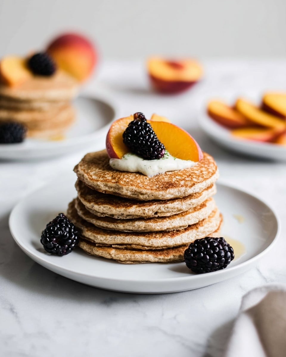 A stack of five light brown pancakes sits in the center of a white plate on a white marbled surface. The pancakes have a soft, slightly rough texture and are topped with a thin layer of cream, two fresh blackberries, and a slice of peach with deep orange and yellow tones. Three more blackberries rest on the plate near the base of the pancake stack. In the blurred background, there is another white plate with pancakes and some peach slices. Photo taken with an iphone --ar 4:5 --v 7