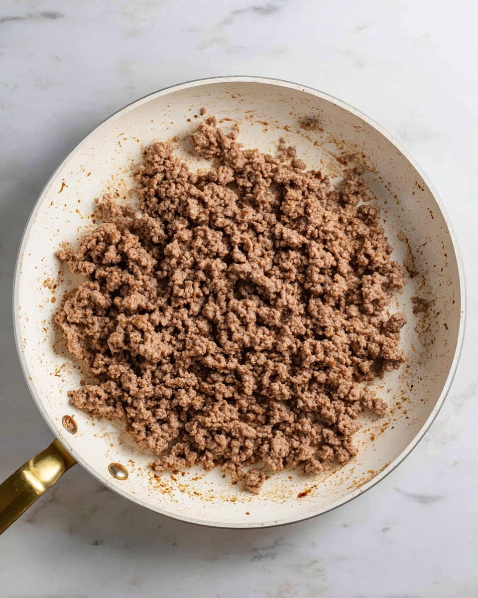 The image shows a white frying pan filled with cooked ground meat unevenly spread out with small and medium chunks, mostly brown with a slightly crumbly texture. The pan's edges have some browned spots and oil stains, and the handle is gold-colored. The frying pan sits on a white marbled surface, visible around the pan. No other ingredients or layers are shown in the image photo taken with an iphone --ar 4:5 --v 7