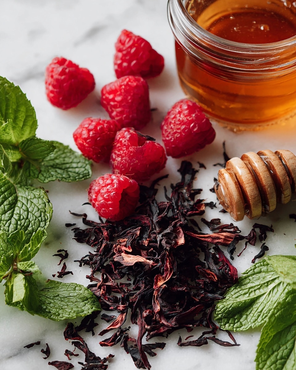 The image shows a close-up of several fresh red raspberries scattered near a small pile of dried dark hibiscus petals on a white marbled surface. A clear glass jar filled with amber honey is placed to the right, with a wooden honey dipper resting inside it. Bright green fresh mint sprigs lie in the bottom left corner, adding contrast with their textured leaves. The arrangement is simple and natural, with the vibrant reds, deep purples, and fresh green colors standing out against the clean white marbled background. photo taken with an iphone --ar 4:5 --v 7