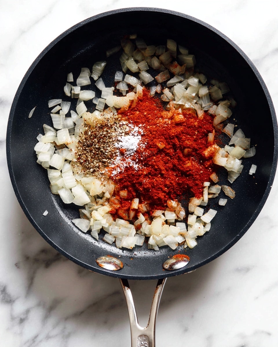 A black pan with a silver handle sits on a white marbled surface, filled with a layer of small, translucent chopped onions scattered evenly across the bottom. On top of the onions, there is a pile of bright red and brown spices, with a sprinkling of white salt, sitting near the center of the pan. The spices form a mostly solid heap with some powdered texture, contrasting with the glossy, slightly cooked onions below. Photo taken with an iphone --ar 4:5 --v 7