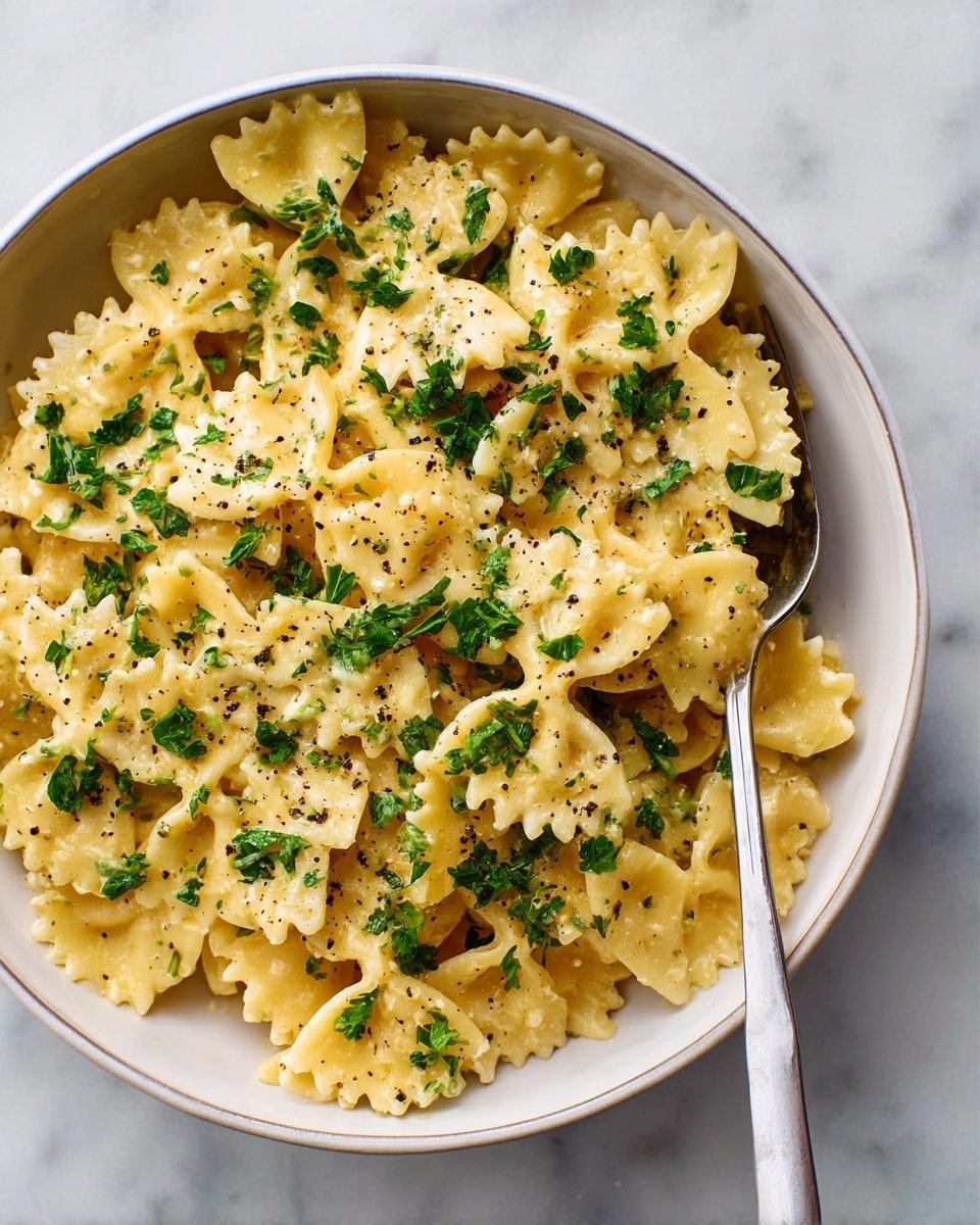 The image shows a white bowl filled with creamy yellow farfalle pasta, covered in a smooth cheese sauce. The pasta has soft edges and is sprinkled generously with bright green chopped parsley and black pepper bits on top. On the right side of the bowl rests a silver fork that curves gently along the bowl’s edge. The bowl is placed on a surface with a white marbled texture. photo taken with an iphone --ar 4:5 --v 7