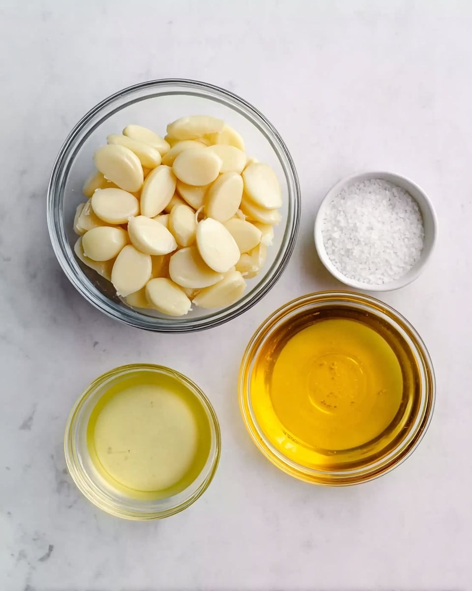 The image shows four clear glass bowls on a white marbled surface. The largest bowl is filled with peeled garlic cloves, which are creamy white and smooth. To its right, there is a bowl of golden yellow oil with a shiny surface. Below the garlic bowl, a smaller bowl contains a light yellow liquid, likely lemon juice. To the right of the lemon juice bowl, a small white bowl holds a coarse white salt. The bowls are neatly arranged, creating a simple, clean layout. Photo taken with an iphone --ar 4:5 --v 7