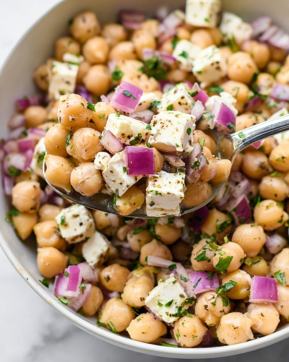 A close-up view of a chickpea salad in a white bowl on a white marbled surface, showing three main layers: beige chickpeas forming the base, mixed with small bright purple pieces of red onion, and topped with white cubes of feta cheese scattered throughout, speckled with finely chopped green herbs and a light sprinkle of black pepper; a silver spoon lifting a mix of chickpeas, onion, and feta from the bowl, highlighting the fresh, textured ingredients. Photo taken with an iphone --ar 4:5 --v 7
