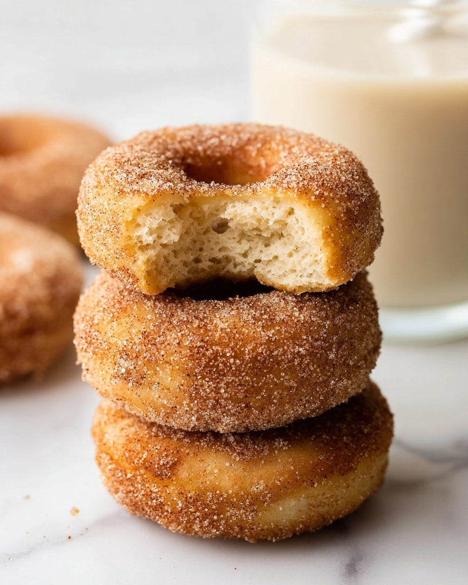 A stack of three cinnamon sugar donuts is shown on a white marbled surface; the top donut has a bite taken from its side, revealing a soft, light beige inside with a slightly crumbly texture, while the outer sides of all donuts are golden brown, evenly coated with granular cinnamon sugar. The donuts are round with a central hole and have a slightly rough surface due to the sugar crystals. In the blurred background, there is a large glass of light beige milk with a white straw. photo taken with an iphone --ar 4:5 --v 7