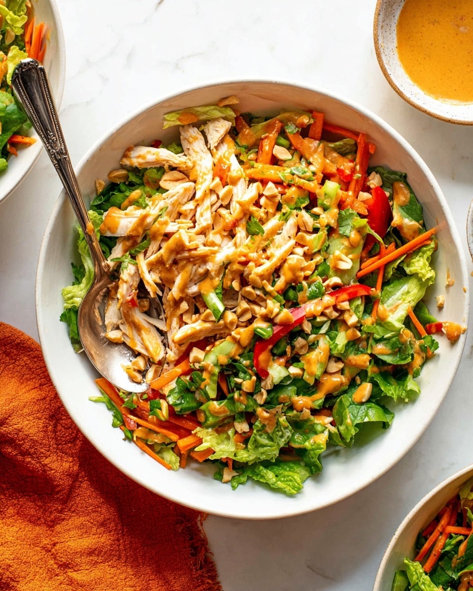 A white bowl filled with a colorful salad featuring three main layers: the bottom layer has mixed green lettuce and chopped cilantro with a fresh green texture; the middle layer contains thin strips of orange carrots, red bell peppers, and green onions; the top layer has shredded white chicken pieces, crunchy peanuts, and is drizzled with a creamy orange-colored dressing. A shiny silver fork and spoon rest in the bowl, slightly buried in the salad. The bowl is placed on a white marbled surface, with a small part of another similar bowl with salad visible at the top right corner. A small dish with leftover dressing is partially seen on the left with an orange cloth nearby. photo taken with an iphone --ar 4:5 --v 7