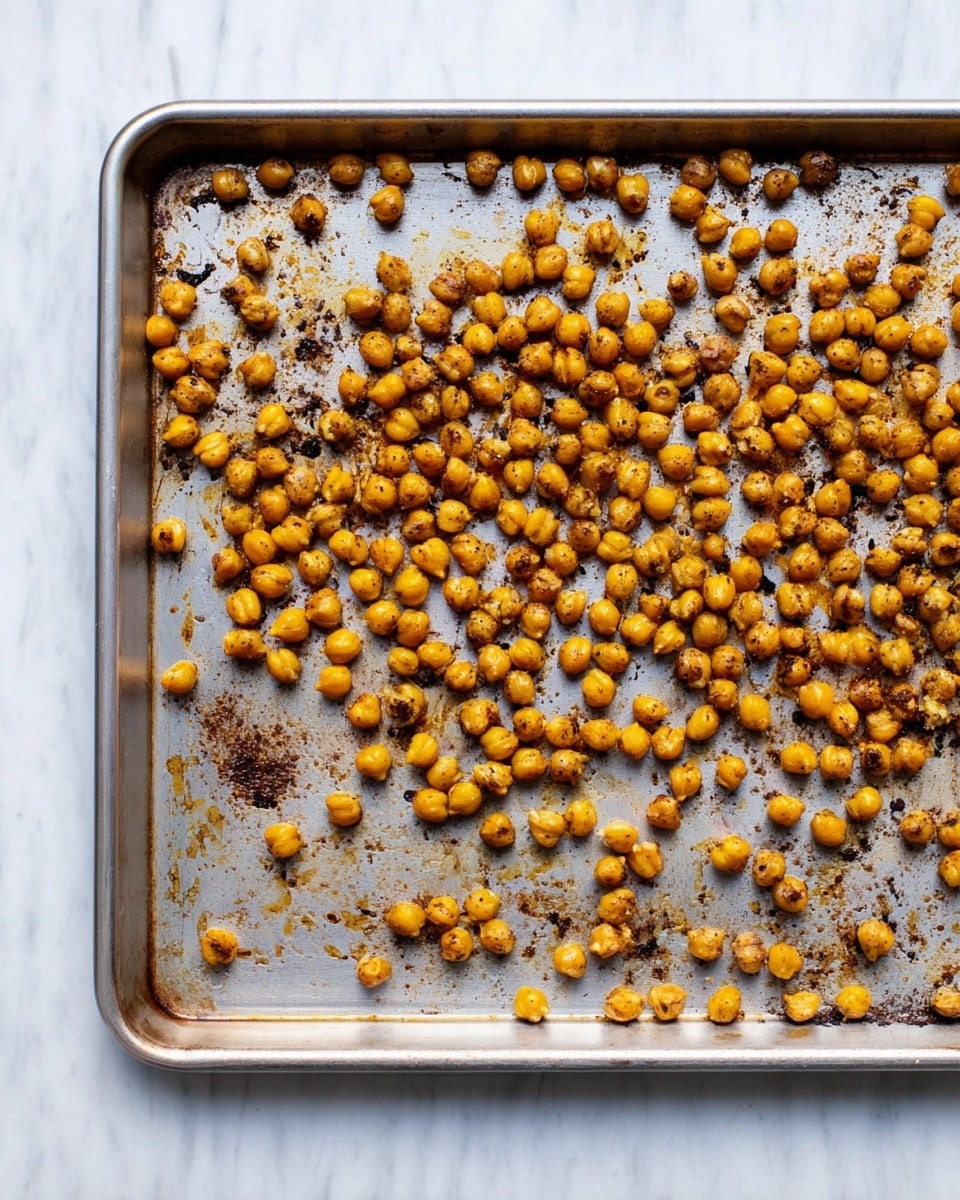 A silver baking tray filled with a single layer of small, round roasted chickpeas that are golden brown with some darker spots. The tray shows some brown baked-on marks from seasoning and roasting. The tray is set on a white marbled surface, giving a clean and bright background. photo taken with an iphone --ar 4:5 --v 7