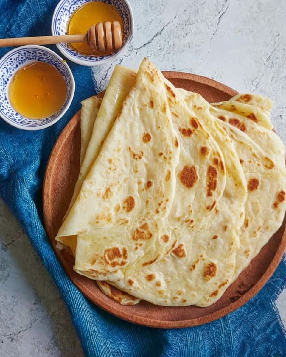 The image shows a round wooden tray placed on a white marbled surface covered with a blue cloth. On the tray, there are four layers of folded flatbread that are light golden with darker brown spots from cooking. The flatbread looks soft and slightly puffy, with some folds overlapping each other. On the top left corner of the image, two small white bowls with blue patterns are placed, one filled with honey which has a shiny and smooth texture. A wooden honey dipper with holes in it lies nearby. The whole setting gives a warm and inviting feeling. photo taken with an iphone --ar 4:5 --v 7