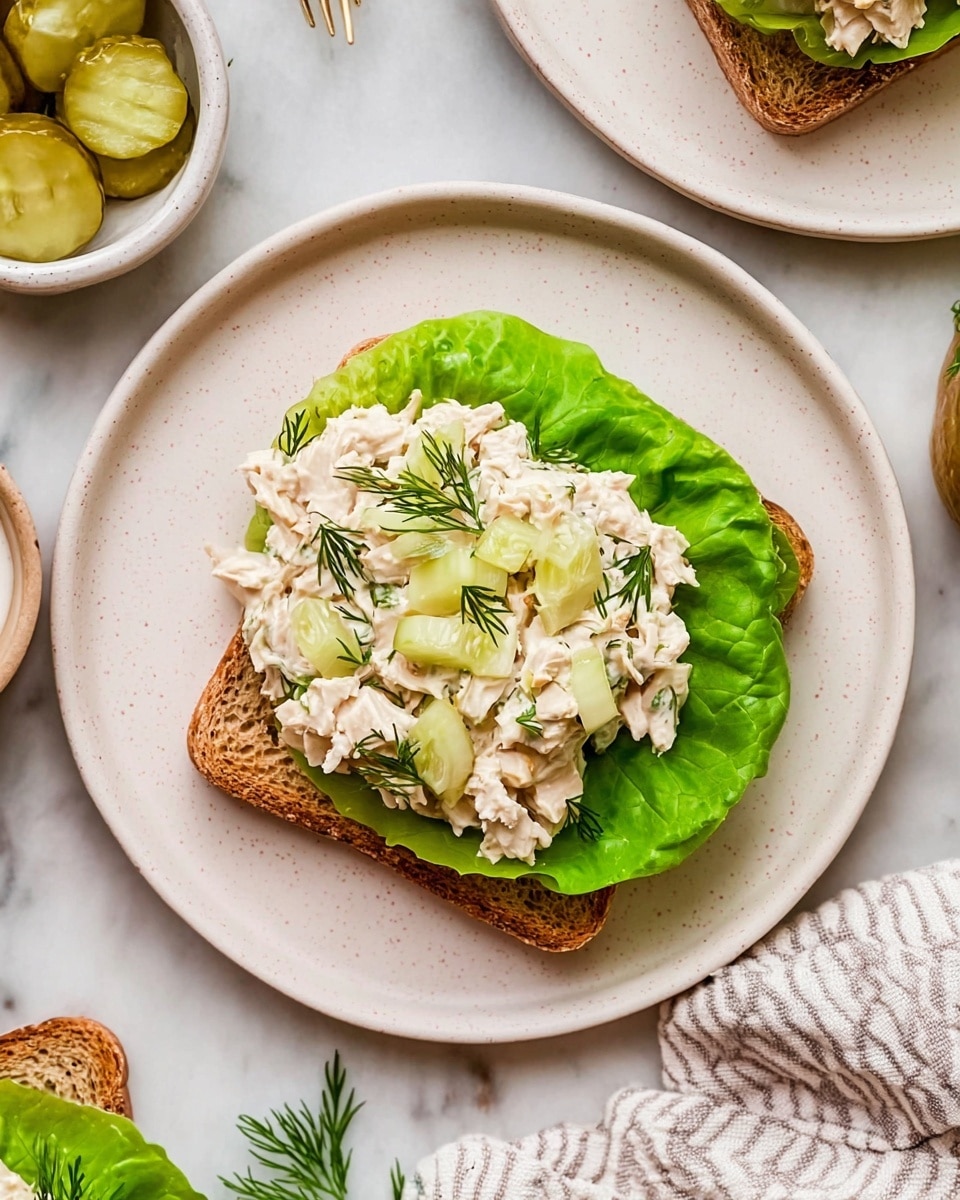 A white plate on a white marbled surface holds an open sandwich made of one slice of toasted brown bread. On the bread lies a bright green lettuce leaf as the first layer, topped with a creamy chicken salad mixed with pale green pickle pieces and finely chopped onions. Several pale green pickle slices and small sprigs of fresh dill rest on top, adding texture and color contrast. Around the plate, there are small bowls with pickle slices and a white sauce, with a soft striped cloth nearby. Photo taken with an iphone --ar 4:5 --v 7