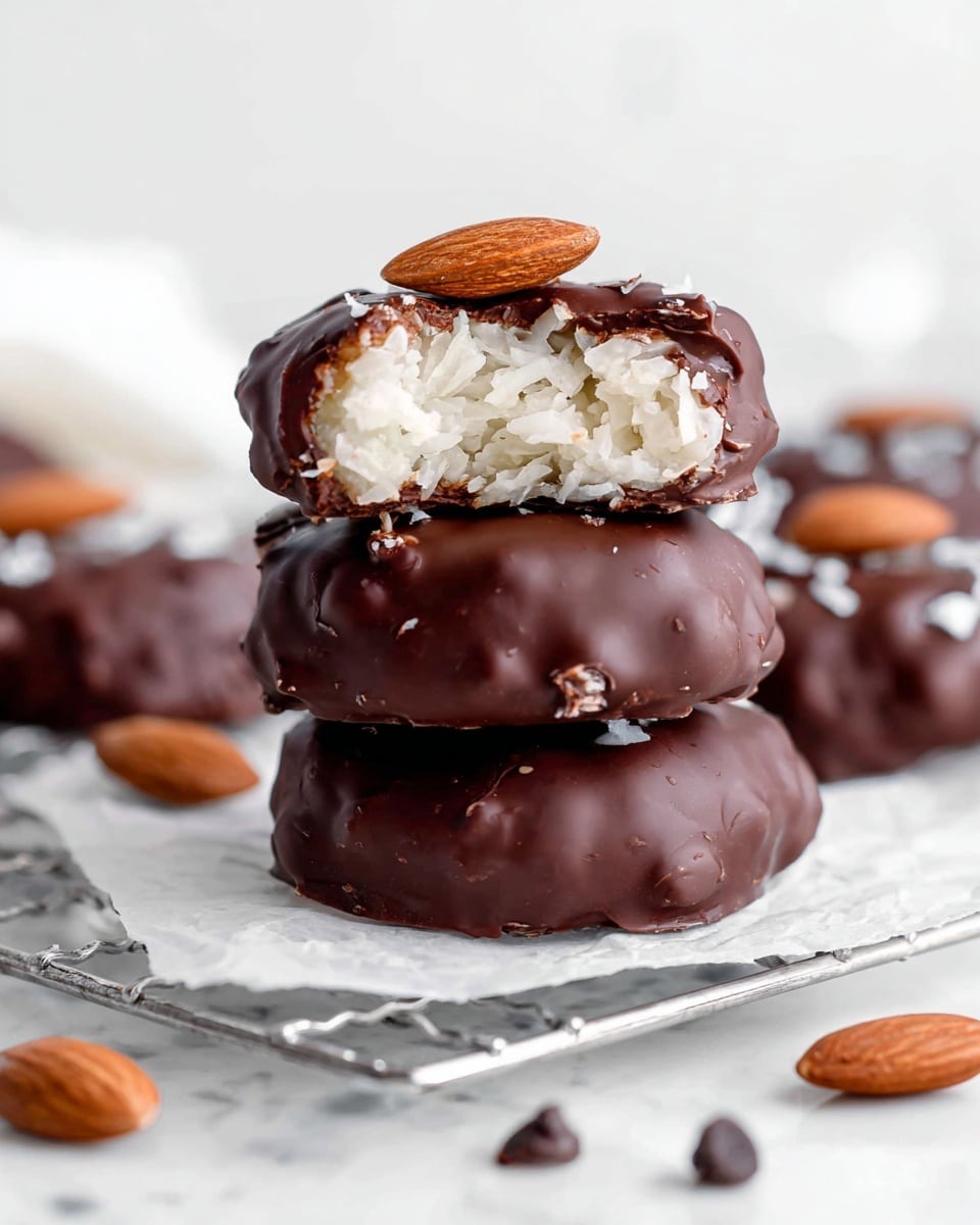 The image shows a close-up of a stack of three chocolate-covered treats on a white marbled surface with a wire rack below. The bottom two treats are fully covered in glossy dark brown chocolate with a slightly rough texture, and the top treat is bitten open, revealing a white, shredded coconut filling inside with a smooth milk chocolate coating around it. A single almond nut sits on top of the bitten treat. Around the stack, there are a few whole almonds and chocolate chips scattered on white parchment paper. The background is bright and white, keeping focus on the treats. Photo taken with an iphone --ar 4:5 --v 7