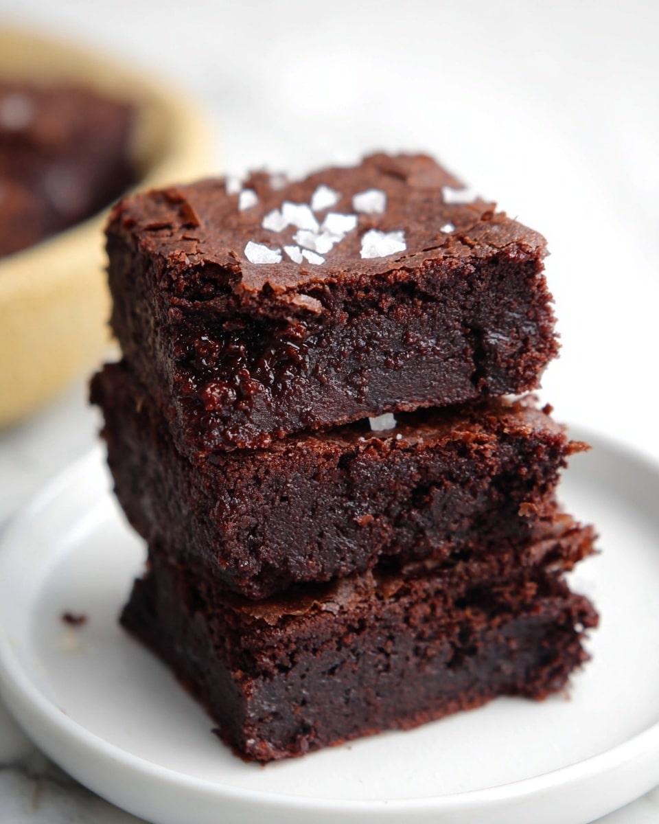 A stack of three dark brown brownies is shown on a white plate. Each brownie has a rich, dense texture with small cracks on the surface. The top brownie has a few white salt flakes scattered on it. The plate sits on a white marbled surface with a blurred background in soft light. photo taken with an iphone --ar 4:5 --v 7