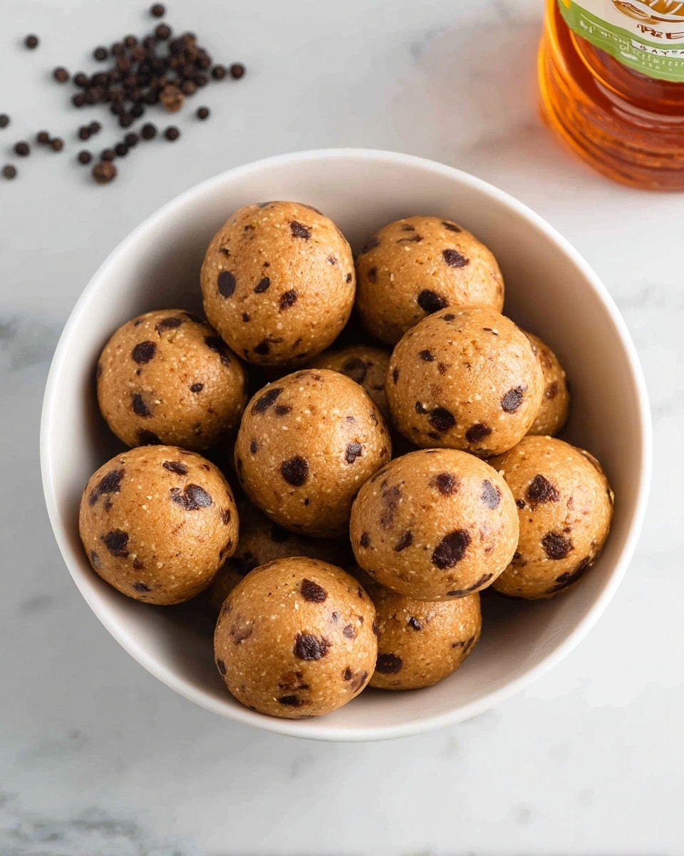 The image shows a white bowl filled with small round energy balls. Each ball is light brown in color, smooth but with visible small dark chocolate chips speckled throughout. The balls are tightly packed in the bowl, which sits on a white marbled surface. In the top right corner, there is a bottle of honey partially visible, and in the top left corner, some scattered black peppercorns add contrast to the clean background. Photo taken with an iphone --ar 4:5 --v 7