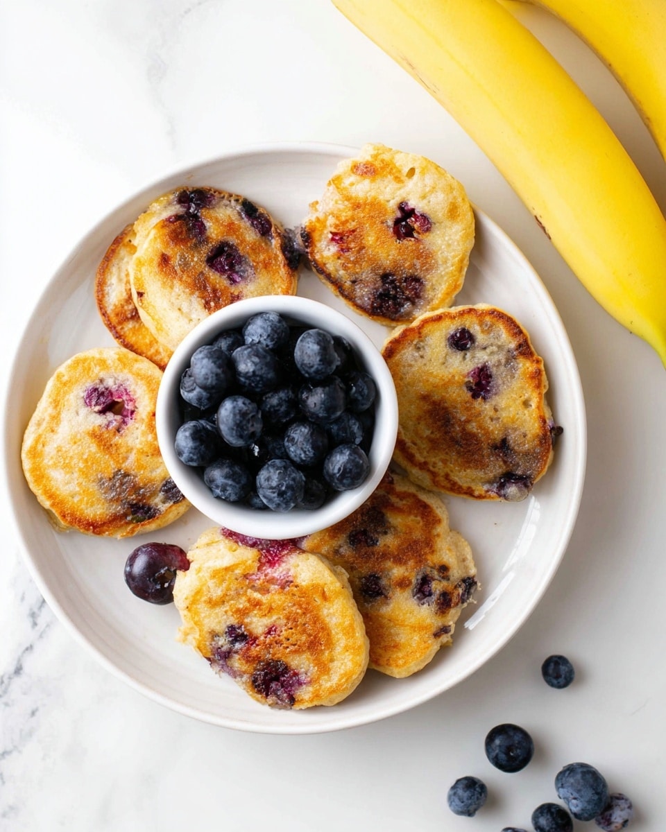The image shows a white plate with six small golden brown pancakes arranged in a circle around a small white cup full of fresh blueberries. The pancakes have visible blueberries baked inside them, appearing as small purple spots with a slightly crispy texture. There is a yellow banana placed in the top right corner on the white marbled surface, and a few loose blueberries scattered near the bottom right of the plate. The overall setting is bright with a clean, white marbled background. photo taken with an iphone --ar 4:5 --v 7