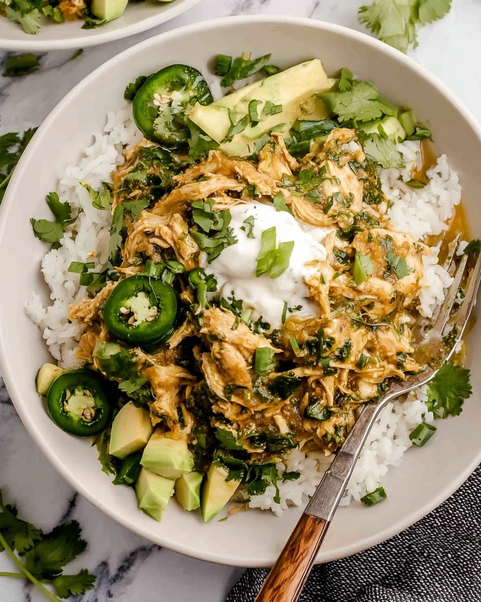 The dish is served in a white bowl on a white marbled surface. The bottom layer is white rice, topped with shredded chicken mixed with green herbs and sauce that has a light brownish-yellow color. There are fresh green cilantro leaves spread around the chicken and rice. Slices of green jalapeño peppers and small cubes of pale green avocado are scattered on top and around the chicken. A dollop of white sour cream sits in the center of the chicken, garnished with a small piece of green onion. A fork with a wooden handle rests on the edge of the bowl. Photo taken with an iphone --ar 4:5 --v 7