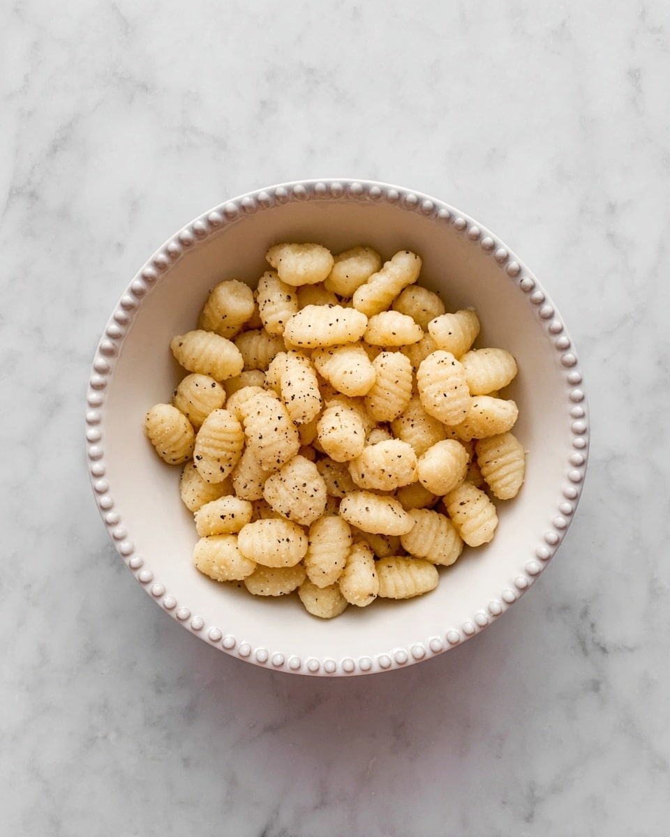 A bowl filled with small, oval-shaped gnocchi pieces that are light golden with a soft, slightly powdery texture, speckled lightly with black pepper. The gnocchi are piled evenly inside a white bowl that has a delicate beaded edge. The bowl sits centered on a white marbled surface, creating a clean and simple background that highlights the pale yellow pasta. photo taken with an iphone --ar 4:5 --v 7