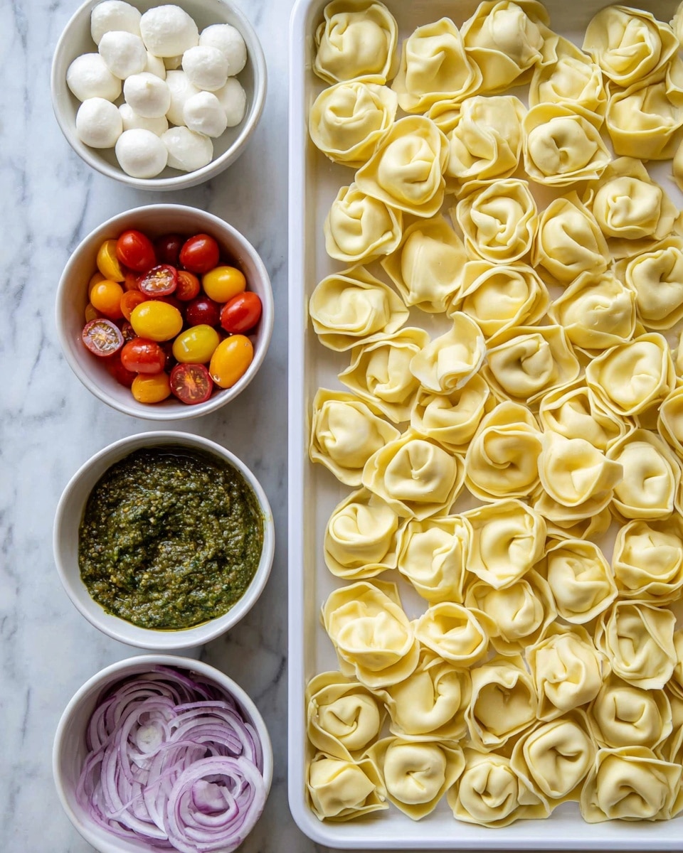 A white tray filled with a single layer of uncooked tortellini, each piece pale yellow and folded into small rings with soft folds and rounded edges, takes up the right side of the image. On the left side, there are four round white bowls placed in a vertical line on a white marbled surface. The top bowl is filled with small white mozzarella balls. Below it, a bowl with thick green pesto sauce showing a chunky texture. Next, a bowl contains colorful halved cherry tomatoes in red, yellow, orange, and purple shades. The bottom bowl holds thinly sliced purple onions with white centers showing. Photo taken with an iphone --ar 4:5 --v 7
