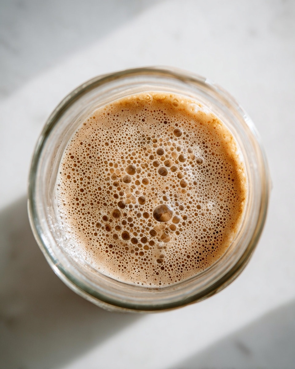 The image shows a close-up top view of a glass cup filled with a light brown frothy liquid. The foam layer on top is dense with many small and medium bubbles, creating a textured surface. The drink fills about half of the transparent cup, placed on a white marbled surface. The glass has smooth edges and the lighting is soft and natural, highlighting the creamy texture of the drink. Photo taken with an iphone --ar 4:5 --v 7