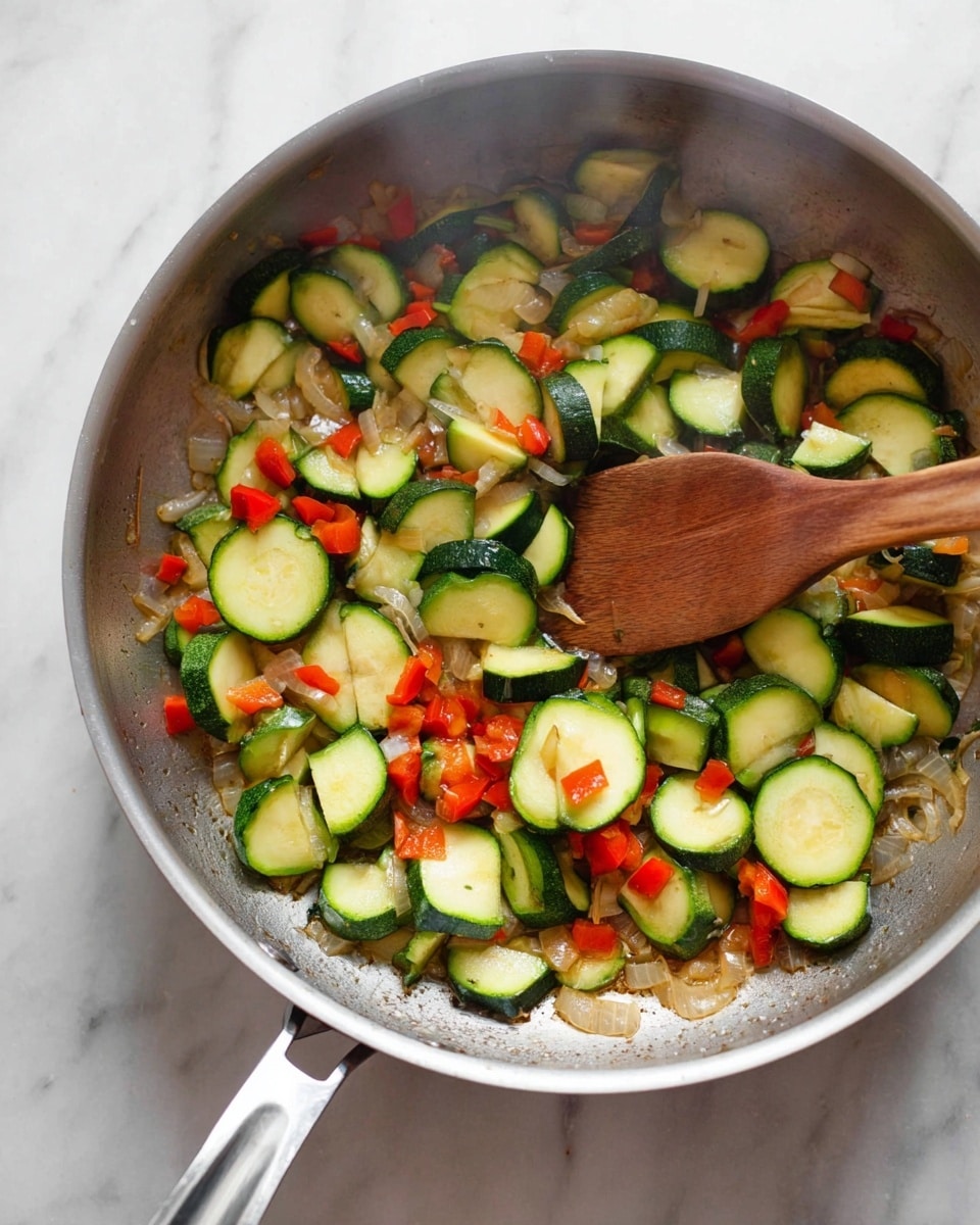 The image shows a silver pan with cooked vegetables, including sliced green zucchini with dark green edges, small red bell pepper pieces, and finely chopped translucent onion. The vegetables are spread evenly across the pan with some light browning on the pan surface. A wooden spoon is partly inside the pan, stirring the vegetables. The pan handle is visible at the bottom, resting on a white marbled surface. steam rises softly from the vegetables. photo taken with an iphone --ar 4:5 --v 7