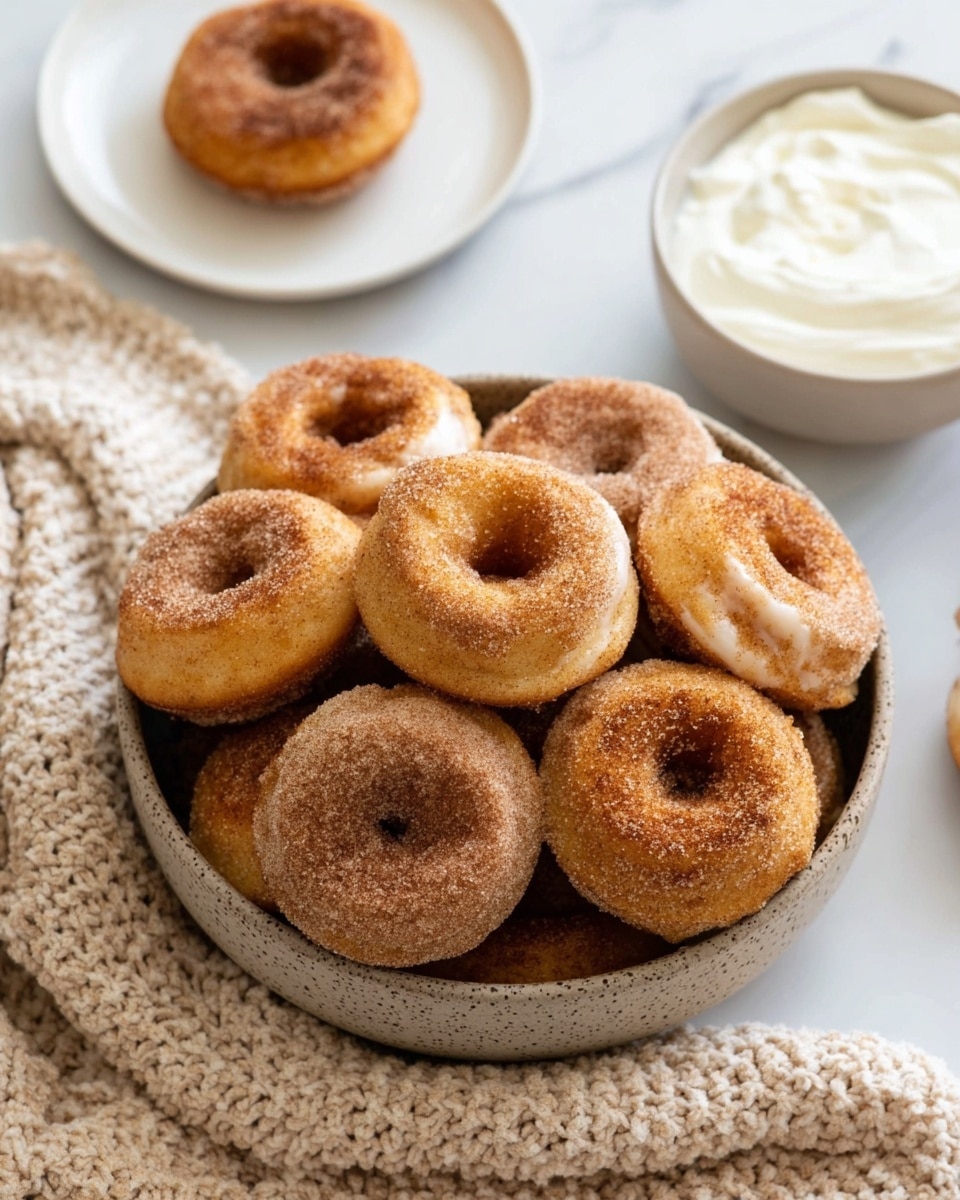A bowl filled with many small round doughnuts, each coated with a layer of cinnamon sugar giving a light brown grainy texture. The doughnuts are stacked closely, showing their soft golden outer surface with a hole in the middle. Next to the bowl is a white bowl of smooth whipped cream and a white plate holding a single doughnut dusted with cinnamon sugar, all placed on a white marbled surface with a beige knitted cloth nearby. The light is natural and soft, highlighting the warm tones of the doughnuts photo taken with an iphone --ar 4:5 --v 7