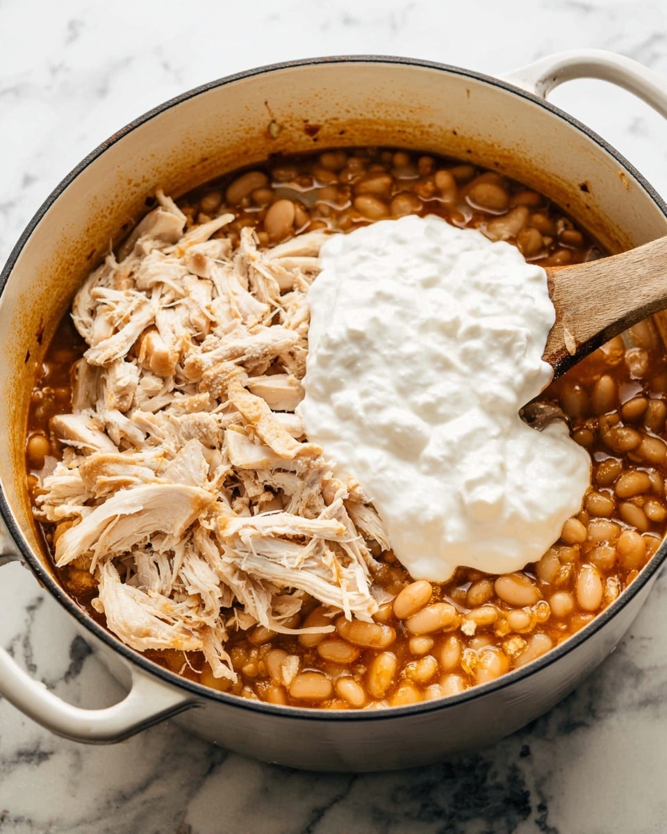 A white pot filled with three visible layers: the bottom layer is a mix of small, soft-beige beans in a reddish-brown sauce; the middle layer consists of shredded light beige chicken pieces scattered evenly on top of the beans; the top layer is a large dollop of thick white creamy sauce covering the middle. Inside the pot, near the edge, there is a wooden spoon partially submerged in the sauce. The pot is placed on a white marbled surface. photo taken with an iphone --ar 4:5 --v 7