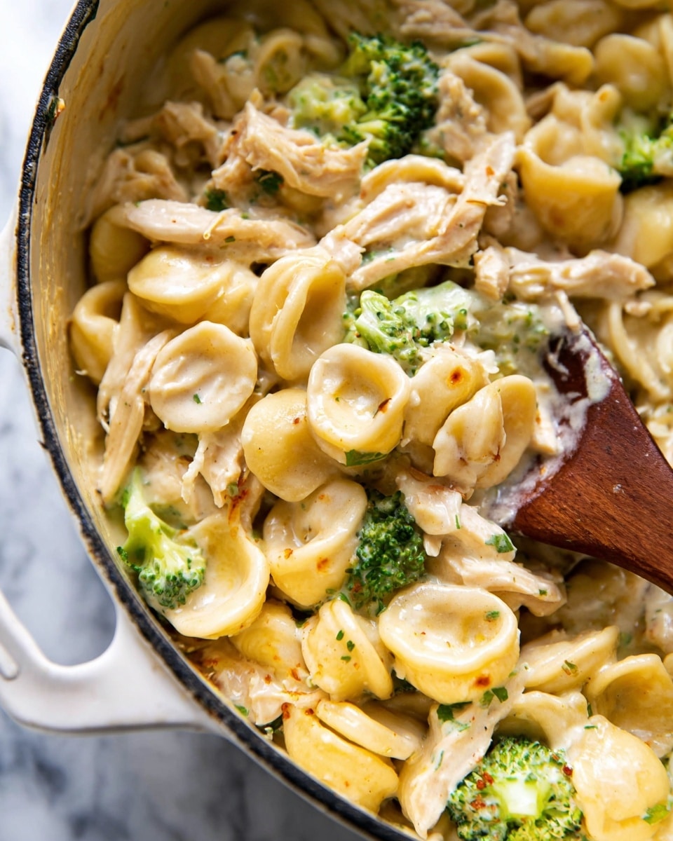 The image shows a close-up of a creamy pasta dish in a white pot with a wooden spoon partially submerged in the creamy sauce. The pasta is orecchiette, light yellow in color with a smooth texture. Mixed in are small, bright green broccoli pieces that add a pop of color. There are shredded pieces of light-colored chicken layered throughout the dish, all coated in a thick, glossy cream sauce that has a slight shine from the light. The background surface is a white marbled texture. photo taken with an iphone --ar 4:5 --v 7