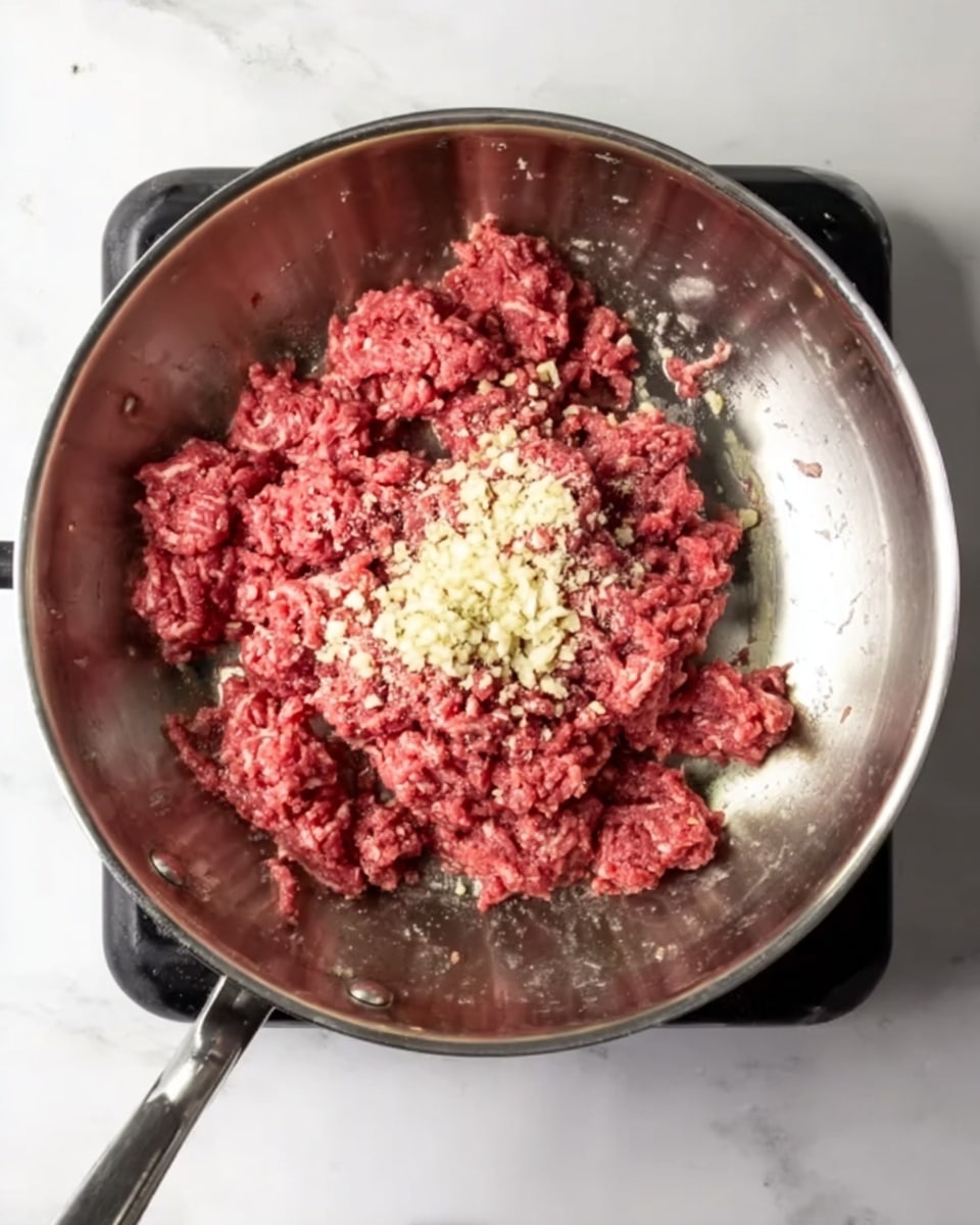 Inside a shiny metal frying pan on a black stovetop, there is a layer of raw ground red meat spread unevenly across the pan's surface. On top of the meat, in the center, there is a small pile of finely minced light beige garlic. The pan is viewed from above on a white marbled surface, with its handle extending out toward the lower left. photo taken with an iphone --ar 4:5 --v 7