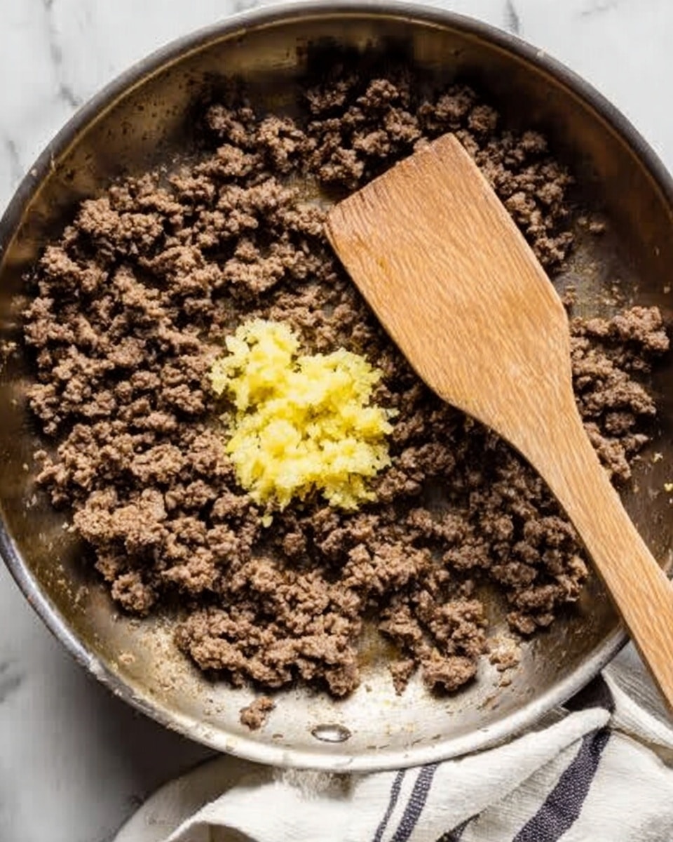 A close-up top view of a skillet filled with cooked ground beef that is brown with a slightly crumbly texture. In the center of the skillet, there is a small pile of finely minced yellow and pale off-white garlic and ginger. A wooden spatula with a smooth texture rests on the right side of the skillet, partially touching the beef. The skillet is on a white marbled surface with a folded white and black striped cloth to the lower right corner. photo taken with an iphone --ar 4:5 --v 7