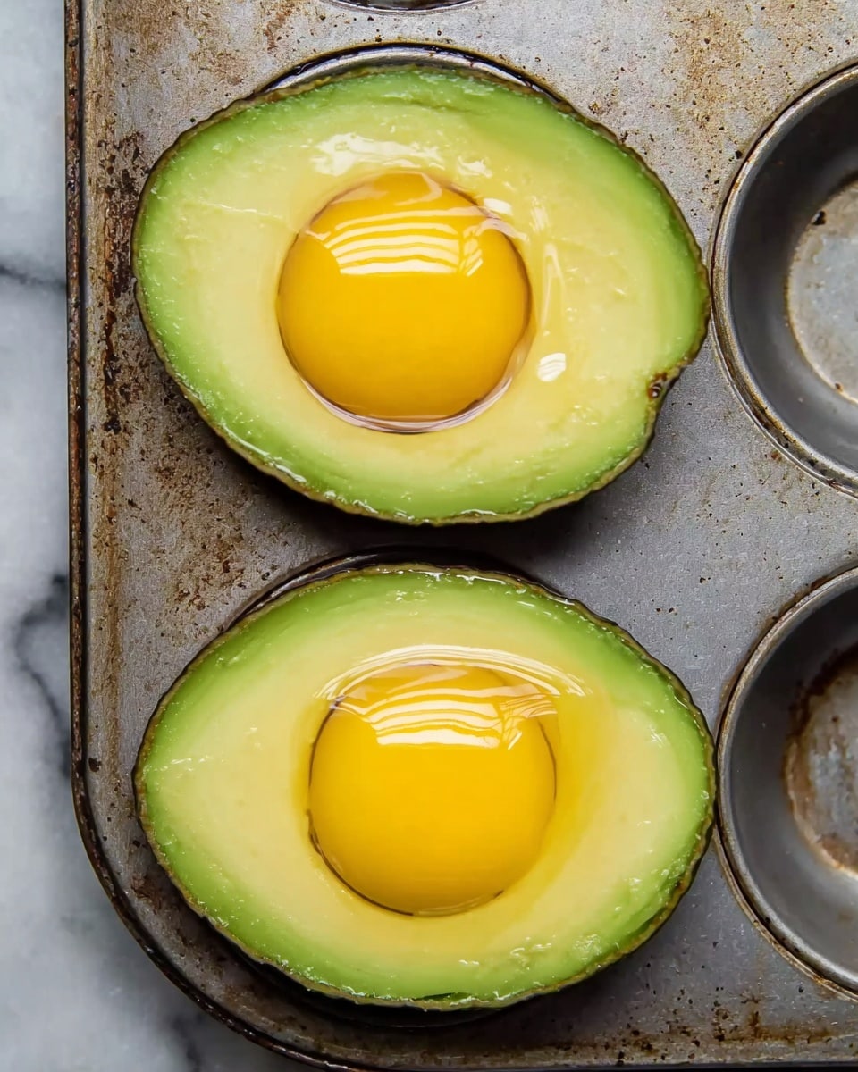 The image shows two avocado halves placed with their rounded edges facing outwards on a muffin tin with a worn metal surface. Each half has a bright green outer skin and a smooth, lighter green inner flesh. In the center of each avocado, a fresh, yellow egg yolk sits smoothly inside the hollow where the seed used to be. The lighting makes the egg yolks shine slightly, and the texture of the metal muffin tin contrasts with the soft avocado. The background has a white marbled texture. photo taken with an iphone --ar 4:5 --v 7
