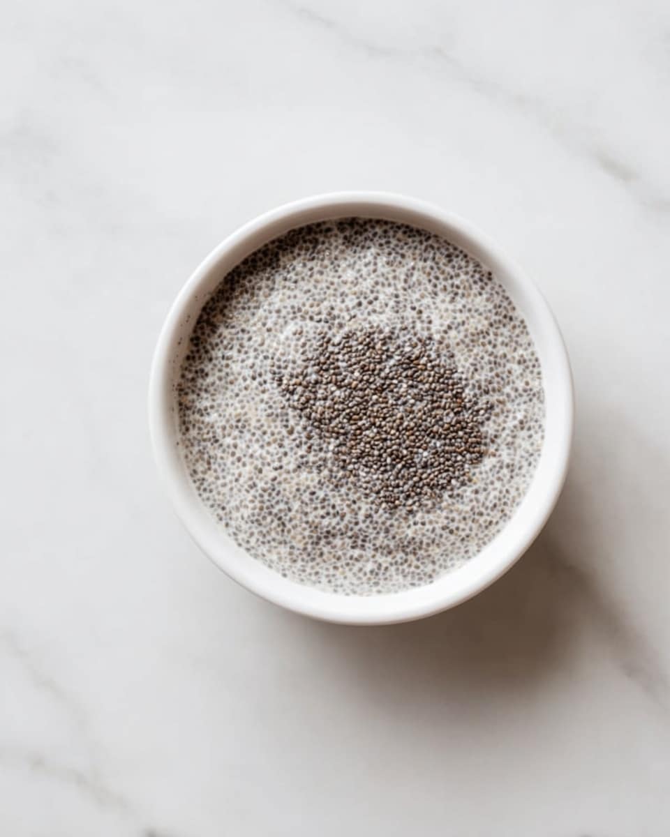 A white bowl filled with a single layer of creamy chia seed pudding, showing tiny dark chia seeds evenly spread throughout the thick, light gray pudding, placed on a smooth white marbled surface, photo taken with an iphone --ar 4:5 --v 7