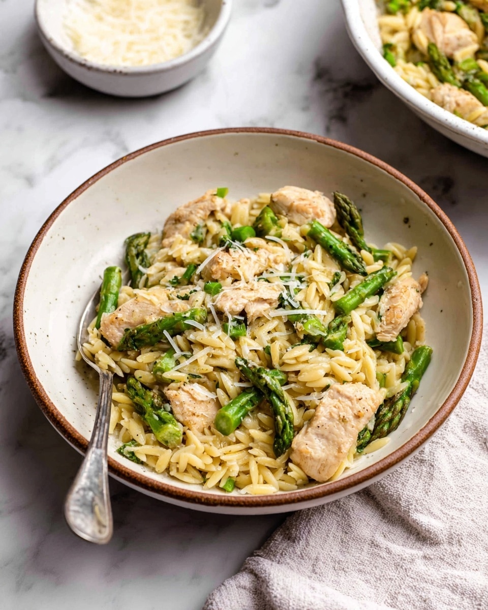 A white bowl with a brown rim holds a dish with three main layers: the bottom layer is small, pale yellow orzo pasta, the middle layer is bite-sized pieces of light golden cooked chicken scattered throughout, and the top layer consists of bright green asparagus pieces mixed evenly. The dish is sprinkled with fine white grated cheese that adds a light texture on top. A silver fork rests inside the bowl on the left side. The bowl sits on a white marbled surface with a small white bowl filled with grated cheese in the background to the upper left and a white dish with the same food to the upper right, next to a soft white cloth. Photo taken with an iphone --ar 4:5 --v 7