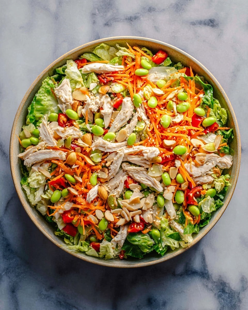A round bowl filled with a colorful salad on a white marbled surface. The bottom layer is green leafy lettuce, topped with shredded orange carrots scattered all over. There are bright red bell pepper pieces mixed inside. Thin strips of white cooked chicken are spread across the salad. Light green edamame beans dot the salad along with sliced light brown almonds and white sesame seeds sprinkled on top. The salad is fresh with varied textures and bright colors. Photo taken with an iphone --ar 4:5 --v 7
