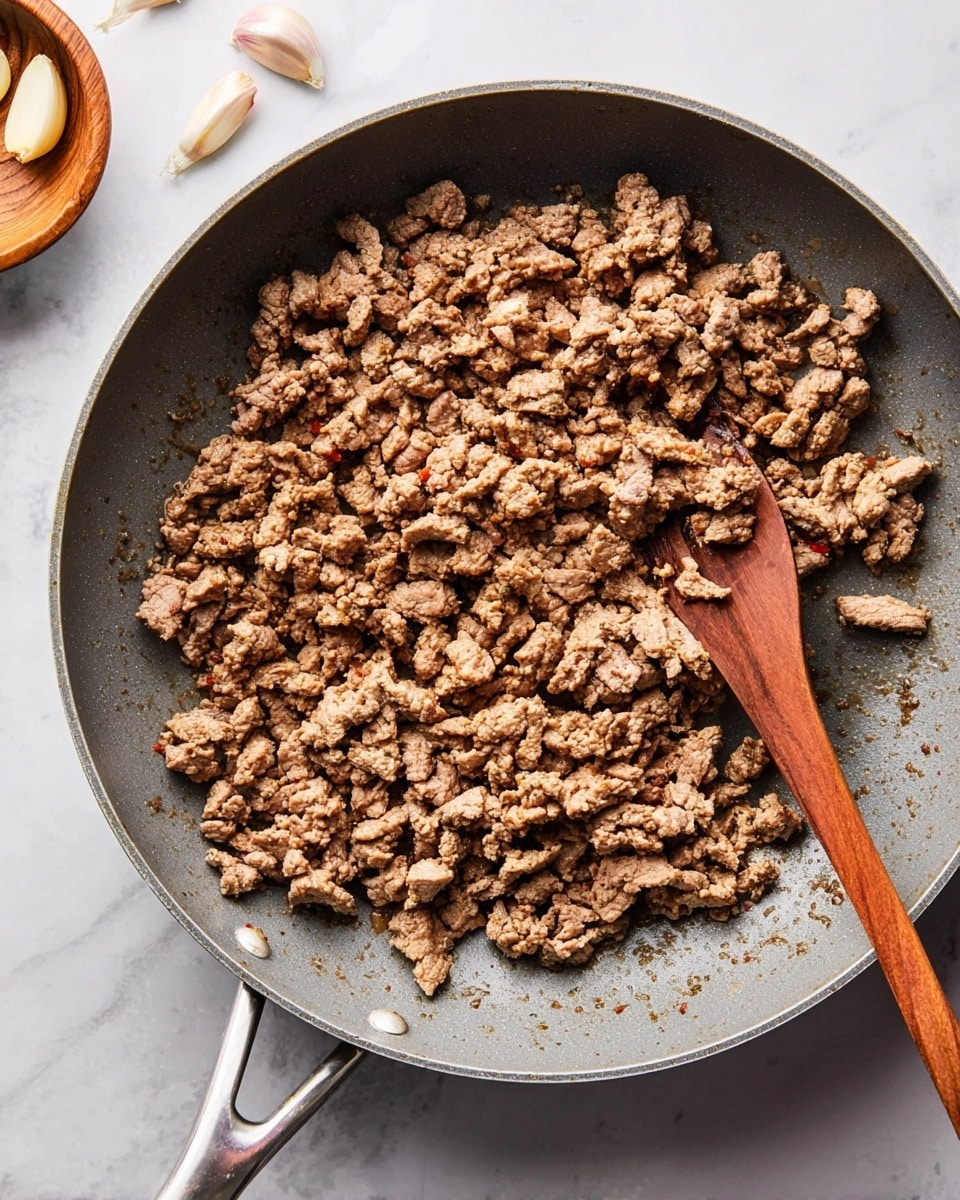 A close-up view of a gray frying pan filled with cooked ground meat, which is browned and crumbly with some small and medium-sized pieces spread evenly across the pan. A wooden spoon with a long handle and darker wood on the scoop side rests on the right side of the pan, partially under the meat. The frying pan has a shiny metal handle and is placed on a white marbled surface. To the upper left, there is a small wooden bowl with some peeled garlic cloves inside. Photo taken with an iphone --ar 4:5 --v 7