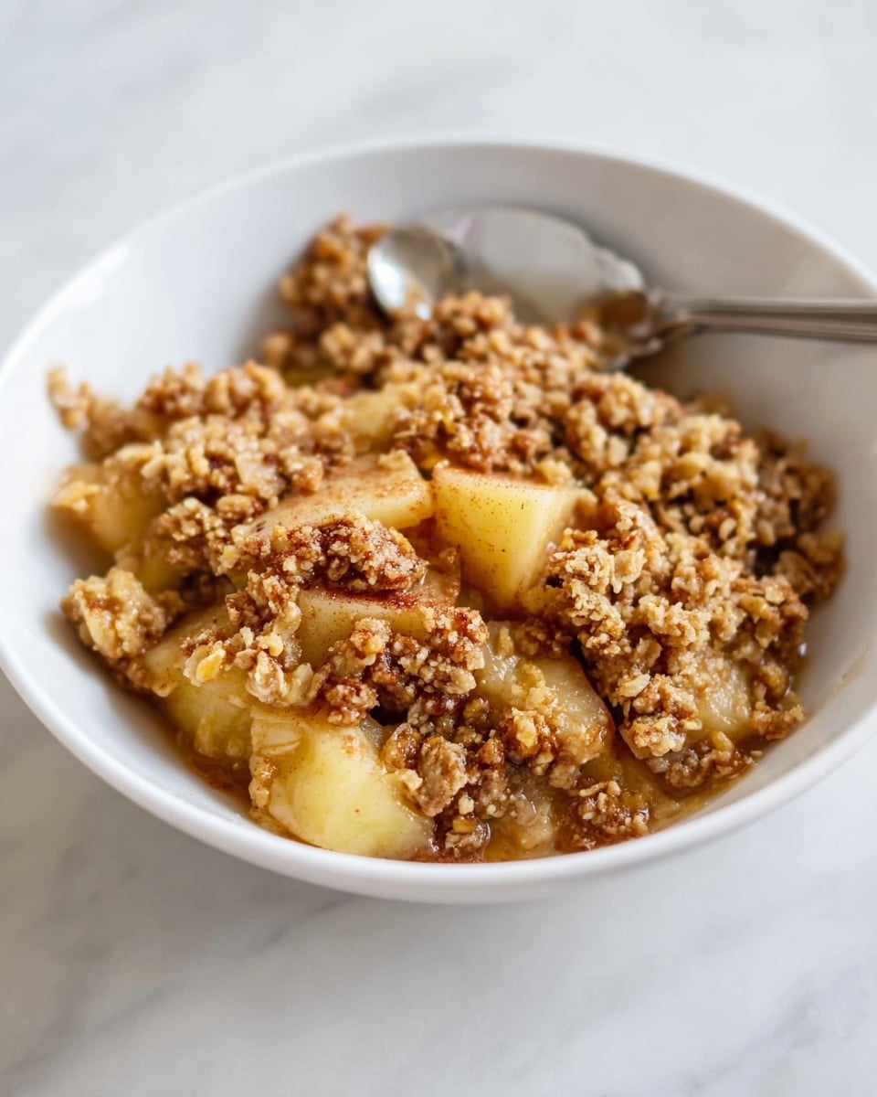 The image shows a white bowl filled with a warm apple crisp dessert. The dish has two main layers: a bottom layer of soft, light yellow cooked apple chunks mixed with brown cinnamon spices, and a top layer of crumbly, golden-brown oat and nut streusel that looks crunchy and textured. A silver spoon is placed inside the bowl, slightly blurred in the background, resting against the side of the bowl. The bowl sits on a white marbled surface, giving a clean and simple setting. photo taken with an iphone --ar 4:5 --v 7