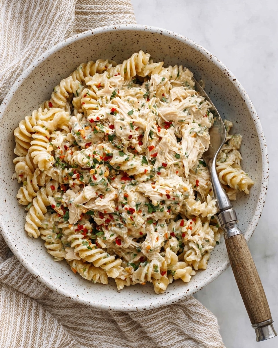 A white speckled bowl holds a creamy pasta dish with two layers visible; the first layer is made of tight spiral rotini pasta in a light beige color, covered by a thick creamy sauce mixed with shredded white meat pieces and small green herb bits scattered throughout. The top is sprinkled with tiny red chili flakes adding a pop of red color, and a silver fork with a wooden handle is placed inside the bowl on the right side. The bowl sits on a white marbled surface with a beige and white striped cloth partially underneath. Photo taken with an iphone --ar 4:5 --v 7