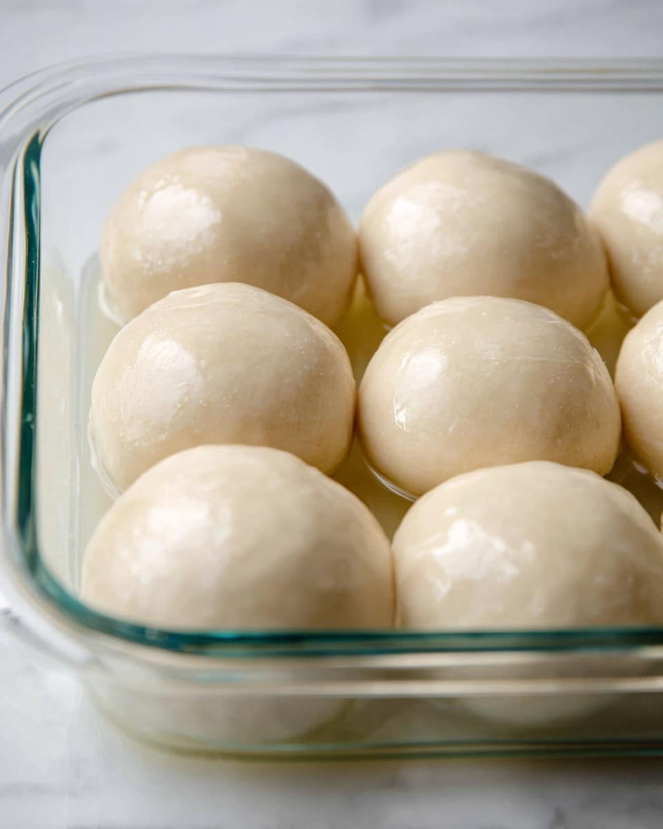 The image shows eight smooth, round dough balls closely placed in a clear glass baking dish. Each dough ball is shiny and pale beige, with a slightly wet look that reflects light softly. The glass dish is set on a white marbled surface, providing a clean and bright background that contrasts with the dough’s subtle color. The dough balls appear soft and plump, resting gently in a thin layer of liquid inside the dish. Photo taken with an iphone --ar 4:5 --v 7