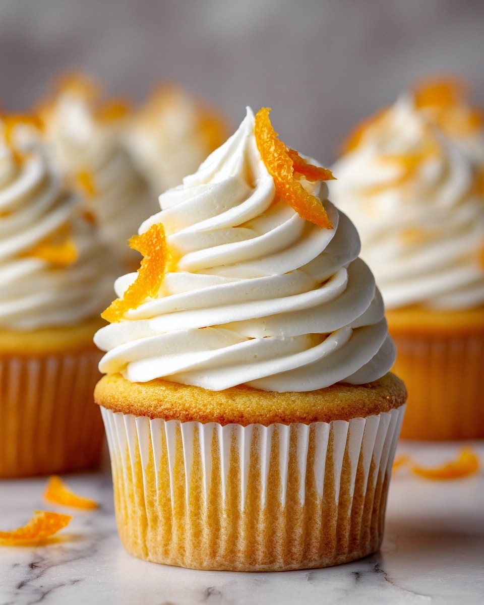 A single cupcake with a golden yellow base wrapped in a white paper liner, topped with three thick, smooth swirls of creamy white frosting stacked on top of each other, and garnished with small pieces of bright orange peel curls sitting on the very top and beside the cupcake. The background shows a soft focus of more similarly decorated cupcakes on a white marbled texture surface. The lighting is warm and natural, highlighting the texture and colors of the cupcake and frosting. photo taken with an iphone --ar 4:5 --v 7