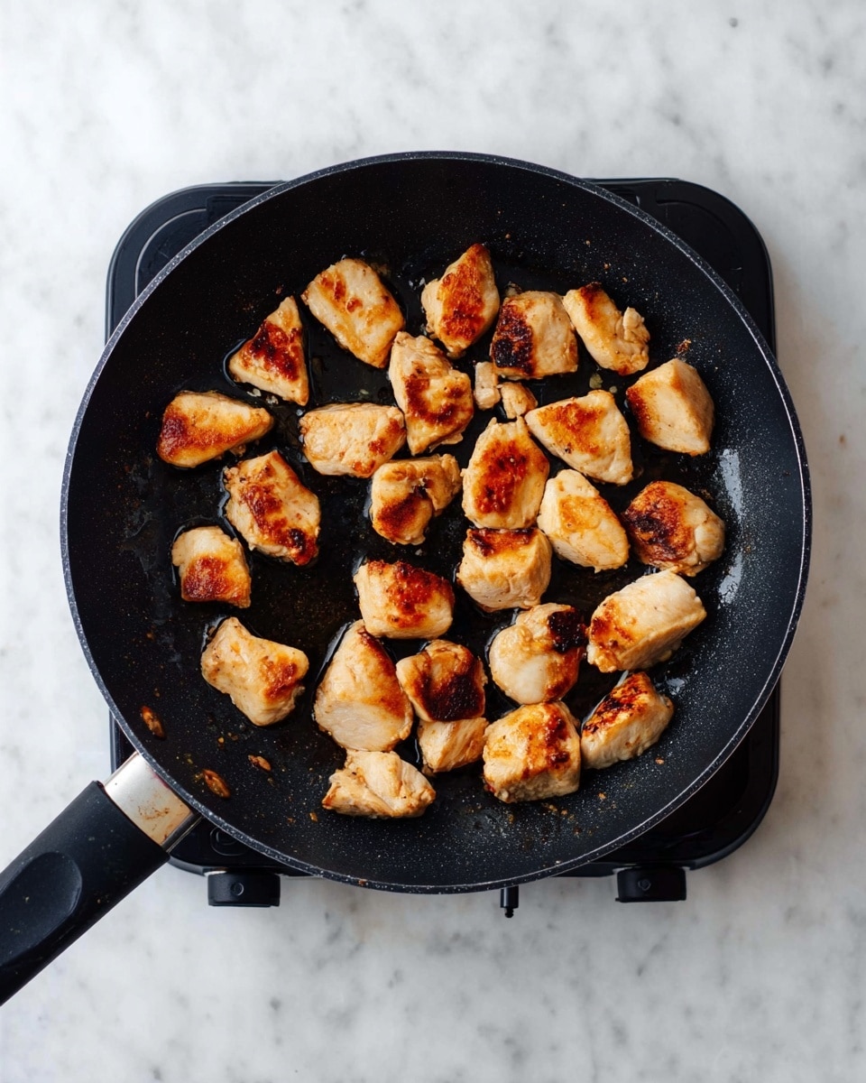 In a black frying pan, there are about twenty small pieces of browned chicken scattered unevenly. Each piece has a golden-brown sear with some darker toasted spots, showcasing a slightly crisp texture. The chicken pieces vary in size and shape, with a moist and cooked surface that gleams slightly from the oil in the pan. The frying pan is placed on an electric burner, and the whole scene is set against a white marbled surface. photo taken with an iphone --ar 4:5 --v 7