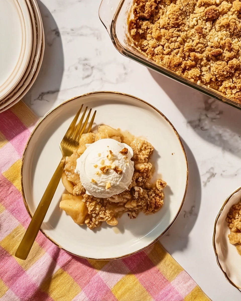 A white plate with a gold rim holds a single serving of a crumbly apple dish topped with a scoop of white ice cream sprinkled with small nut pieces, resting on a white marbled surface. The dish has a thick layer of light golden brown crumble on top with a soft, slightly chunky apple filling beneath, showing a mix of creamy beige and light brown tones. A shiny gold fork lies diagonally across the plate on a pink and yellow checkered cloth, next to a stack of plain white plates with gold rims. In the upper right corner, a clear white baking dish contains the remaining crumbly apple dessert with a textured golden crust and visible chunks, placed on the same white marbled surface. The scene is softly lit, focusing on the textures and colors of the food photo taken with an iphone --ar 4:5 --v 7