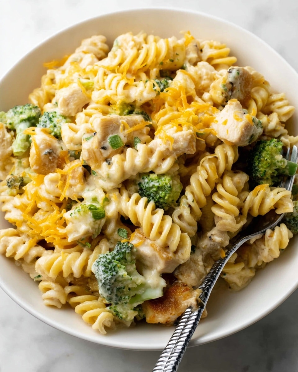 A white bowl filled with rotini pasta coated in a creamy light yellow sauce, mixed with small green broccoli pieces and chunks of light brown cooked chicken. The pasta has a slightly firm texture, with some bits of melted orange cheese scattered on top. There is a silver fork with a black handle resting on the bowl's edge. The bowl sits on a white marbled surface. photo taken with an iphone --ar 4:5 --v 7