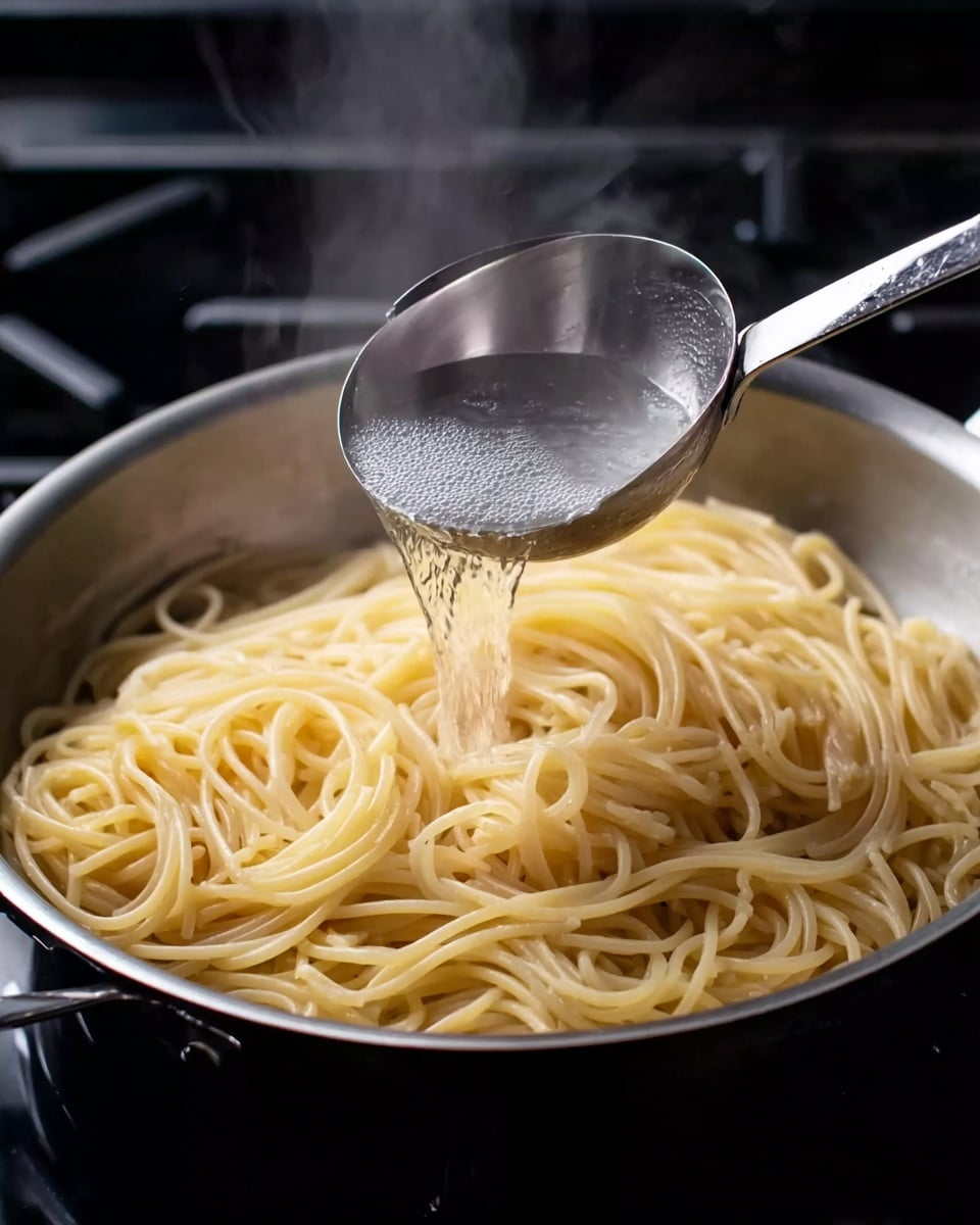 A close-up image shows a pan filled with cooked spaghetti noodles that are pale yellow and slightly shiny, arranged loosely all around the pan. A metal ladle with clear hot water is held above the noodles, pouring water onto them. Steam rises from the pan, giving a warm and fresh cooking feeling. The pan is placed on a stovetop with a black surface visible around it. Photo taken with an iphone --ar 4:5 --v 7