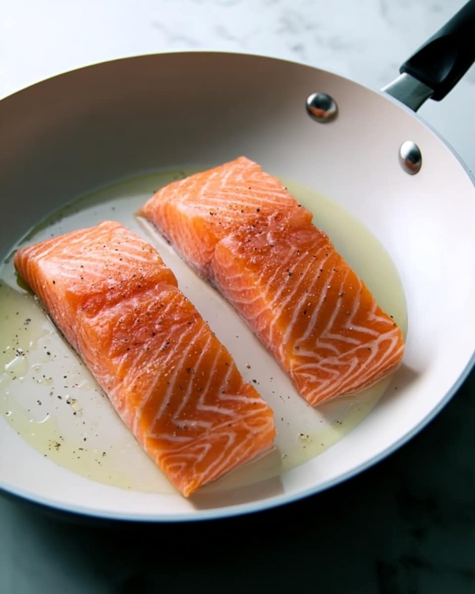 Two pieces of raw salmon with light pink-orange color and white lines are placed side by side in a white frying pan with a black handle. The salmon pieces have some black pepper sprinkled on top, and there is a small amount of clear cooking oil around them. The pan is set on a white marbled surface. photo taken with an iphone --ar 4:5 --v 7