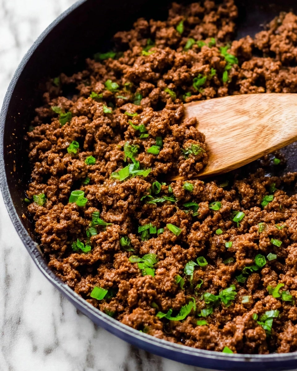 A close-up view of a black round pan filled with cooked ground meat, browned and crumbly with an even texture spread all over. Bright green chopped herbs are scattered across the meat, adding small pops of fresh color. A wooden spoon is scooping some of the meat from the right side of the pan, showing the moist and well-seasoned texture. The pan sits on a white marbled surface. photo taken with an iphone --ar 4:5 --v 7