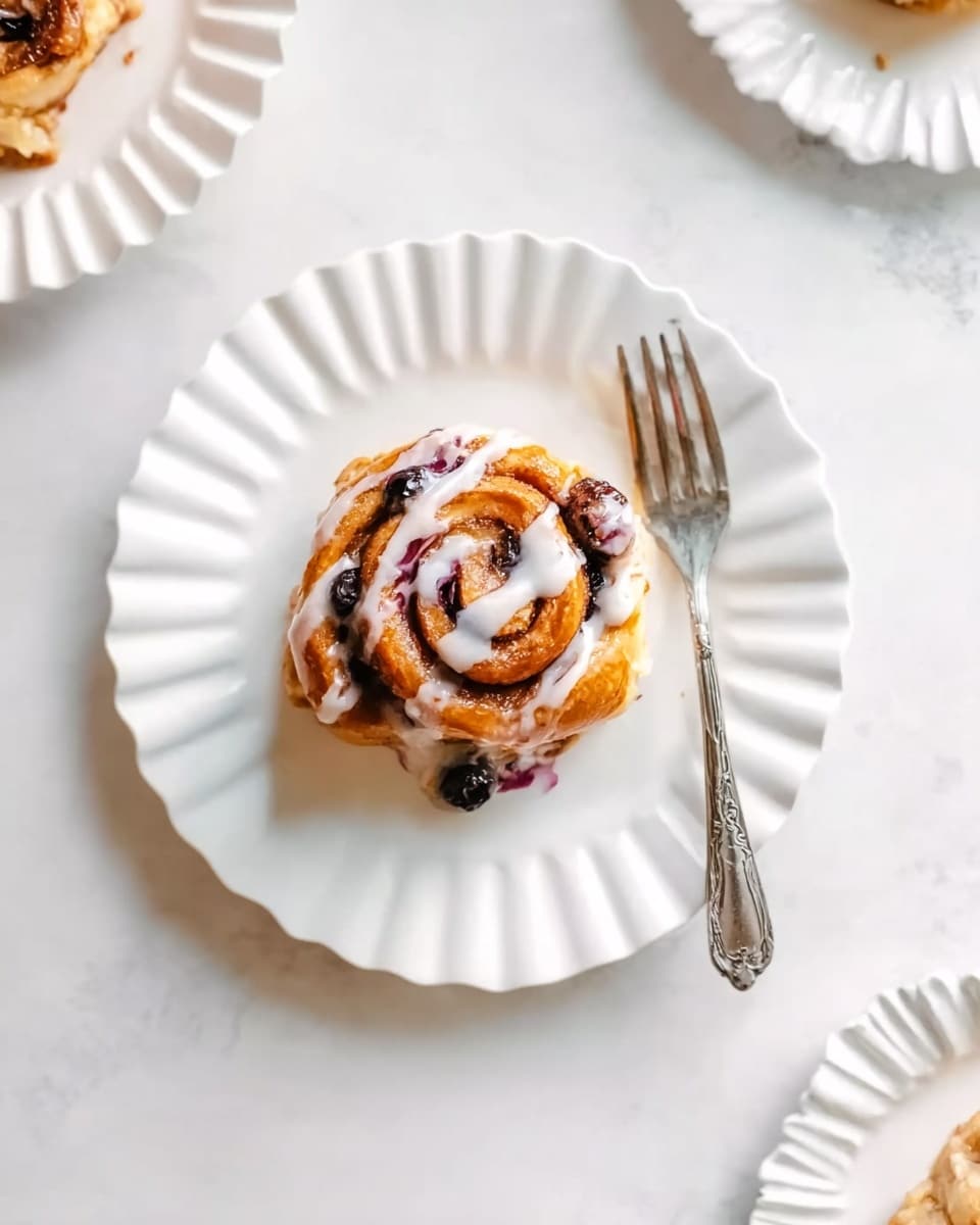 A single cinnamon roll sits in the middle of a white plate with a scalloped edge, topped with white icing drizzled over a golden-brown spiral dough filled with dark berries and cinnamon, with a silver fork resting next to it on the plate; the plate is placed on a white marbled surface, and part of two other white scalloped plates with parts of cinnamon rolls are visible at the edges of the image photo taken with an iphone --ar 4:5 --v 7