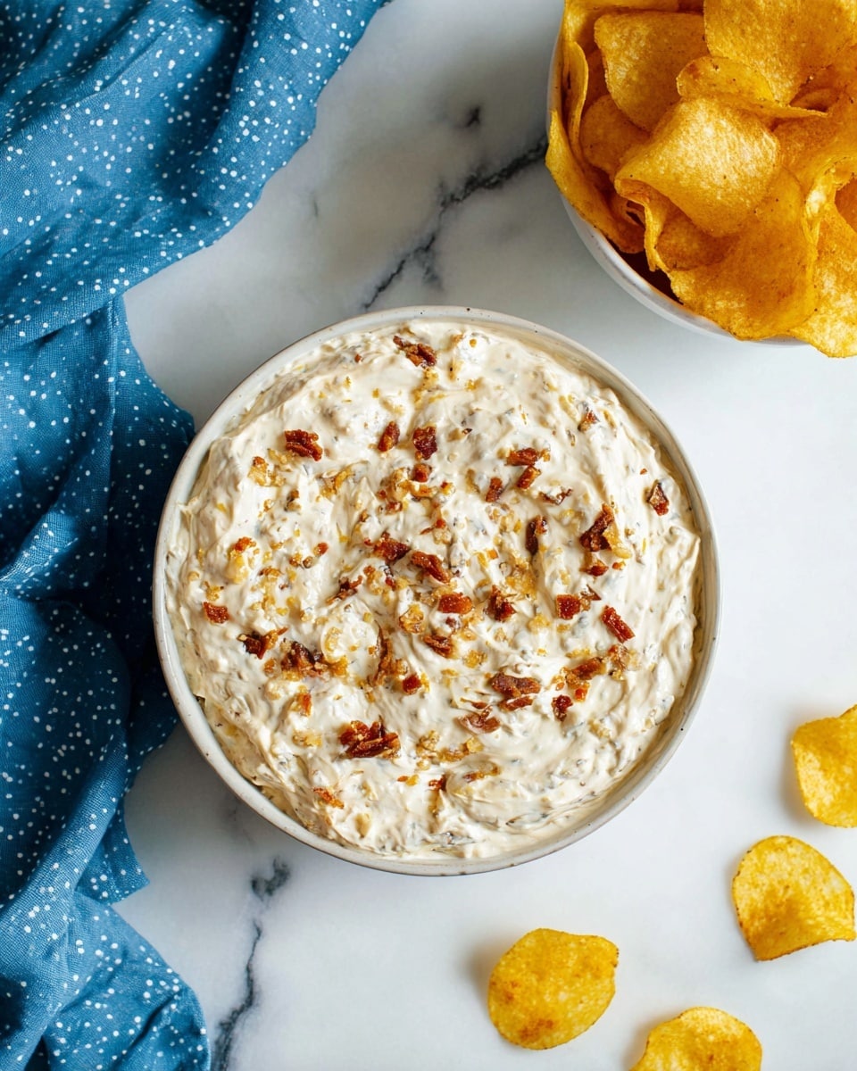 A bowl filled with a creamy white dip mixed with small bits of browned, crispy pieces scattered throughout, giving the dip a textured, uneven surface. The bowl is white and round, placed on a white marbled surface. Next to it, there is a white bowl filled with golden yellow potato chips that have a slightly rough texture. A few potato chips are scattered on the white marbled surface nearby, and a crumpled blue cloth with white spots lies at the bottom left corner. The scene is bright and clean with natural lighting, showing the creamy dip as the main focus photo taken with an iphone --ar 4:5 --v 7