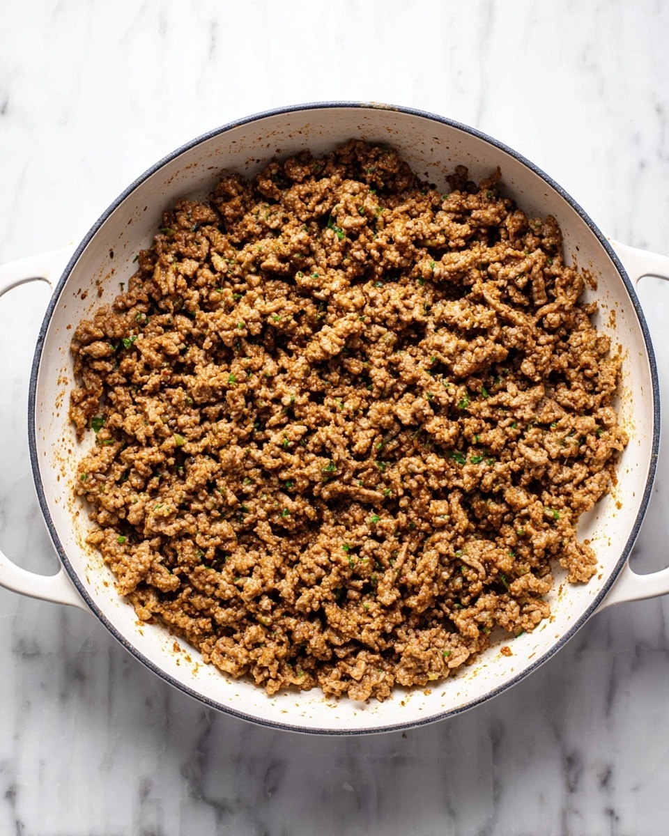 The image shows a white pan filled with cooked ground meat that is brown with small bits of green herbs mixed throughout, giving it a slightly speckled texture. The meat is evenly spread across the pan, and the pan itself has two handles visible on both sides. The background features a white marbled surface. The overall look is simple, warm, and cooked with visible seasoning. photo taken with an iphone --ar 4:5 --v 7