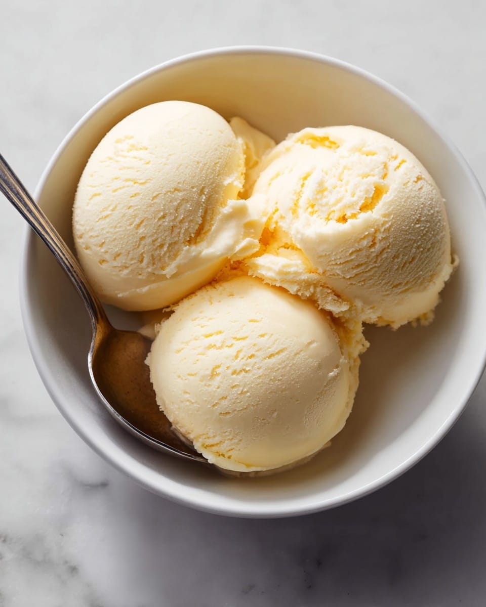 Three smooth, round scoops of pale yellow ice cream sit closely together in a white bowl. The ice cream has a creamy texture with slight roughness near the edges of the scoops. A silver spoon rests inside the bowl next to the ice cream, with the handle extending out and lying on a white marbled surface. The lighting softly highlights the creamy texture and pale color of the ice cream. photo taken with an iphone --ar 4:5 --v 7