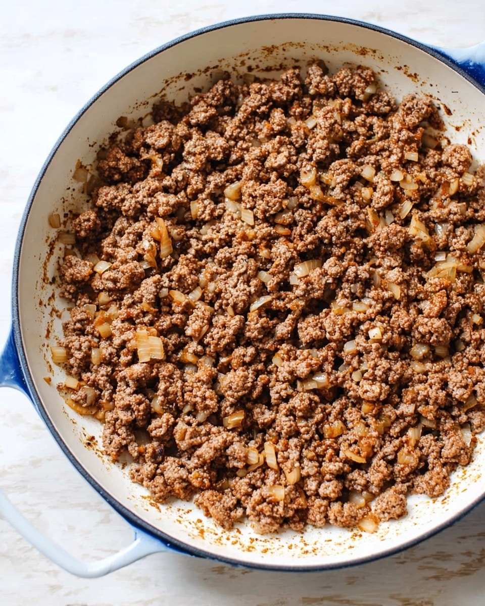 A white skillet with a blue rim is filled with cooked ground meat mixed with small pieces of soft, light brown onions. The mixture covers the entire bottom of the pan, showing a rough, crumbly texture with some browned edges, and a few bits stuck lightly to the sides. The skillet sits on a white marbled surface. photo taken with an iphone --ar 4:5 --v 7