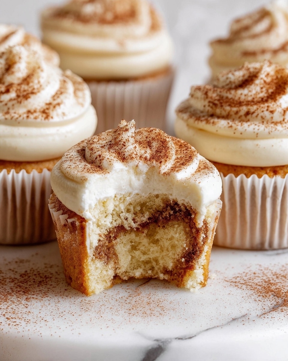 The image shows several cupcakes on a white marbled surface, each with two thick layers of creamy white frosting on top. The frosting is dusted with light brown cocoa powder. One cupcake is bitten, revealing a soft, light yellow cake inside with a darker brown swirl pattern near the outer edge. The cupcakes are in white paper liners, and the frosting on each has a smooth, fluffy texture with a gentle swirl shape. The cocoa powder adds a fine, dusty texture scattered lightly around the cupcakes on the surface. photo taken with an iphone --ar 4:5 --v 7